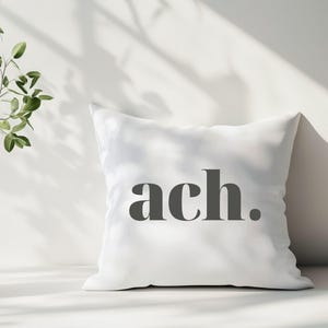 Könnte beinhalten: Ein weißes quadratisches Kissen mit dem Wort "ach." in Dunkelgrau aufgedruckt. Das Kissen steht vor einer weißen Wand, mit Schatten und einer Pflanze im Hintergrund. Ein Einrichtungsgegenstand.