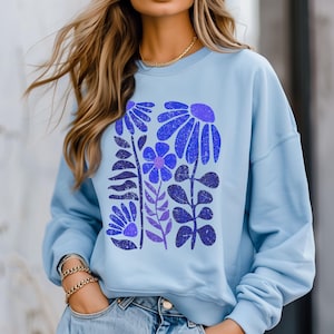 Könnte beinhalten: Hellblaues Sweatshirt mit einem floralen Design in Blau- und Lilatönen. Die Grafik zeigt stilisierte Blumen und Blätter, die ein botanisches Thema bilden. Das Sweatshirt hat einen Rundhalsausschnitt und lange Ärmel.