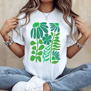 May include: White t-shirt featuring a green floral design. The graphic includes stylized flowers and leaves in varying shades of green. The shirt has short sleeves and a crew neck.