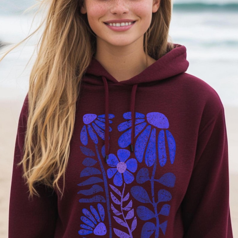 May include: A maroon hooded sweatshirt featuring a graphic print of stylized flowers in shades of blue and purple. The design includes multiple floral stems with leaves and petals, showcasing a distressed texture across the front of the garment.