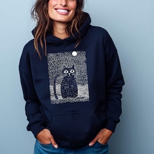 Könnte beinhalten: Marineblauer Kapuzenpullover mit einer Illustration einer schwarzen Katze, einem weißen Mond und einem Sternenhintergrund. Die Katze hat große Augen und einen langen Schwanz. Der Hoodie hat eine Vordertasche und eine Kapuze mit Kordelzug. Ein lässiges, bequemes Kleidungsstück.