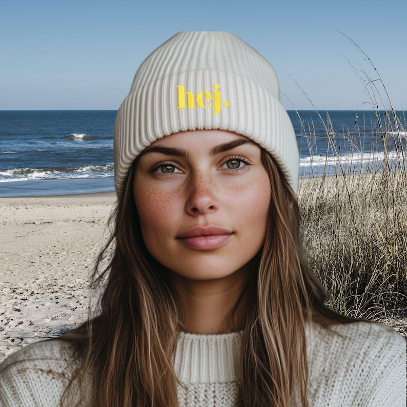 May include: A cream-coloured ribbed knit beanie featuring the word "hej" in yellow. The hat is worn by a person with long brown hair and a cream-coloured sweater. The background shows a beach scene with the sea and tall grass.