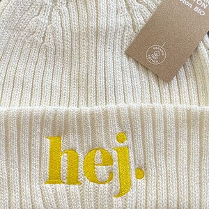 May include: Off-white knitted beanie hat with the word "hej" embroidered in yellow. The hat has a ribbed texture and a folded brim. A brown tag with the words "Organic Cotton Bio" is attached.