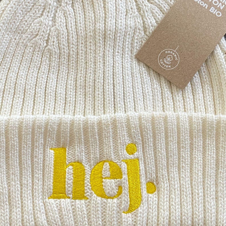 May include: Off-white knitted beanie hat with the word "hej" embroidered in yellow. The hat has a ribbed texture and a folded brim. A brown tag with the words "Organic Cotton Bio" is attached.