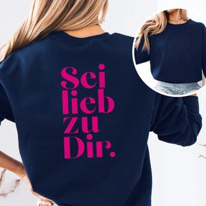 Könnte beinhalten: Ein dunkelblaues Langarm-Sweatshirt mit dem Schriftzug "Sei lieb zu Dir." in fetten, leuchtend pinkfarbenen Buchstaben auf der Rückseite. Die Vorderseite des Sweatshirts ist schlicht dunkelblau, wie im kreisförmigen Inset oben rechts zu sehen.