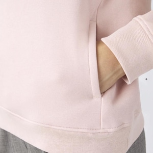 May include: A light pink sweatshirt featuring a smooth, solid-colored fabric, a ribbed cuff on the long sleeve, and a vertical side pocket opening. The garment has a clean, minimalist construction with visible stitching along the hem and pocket edge.