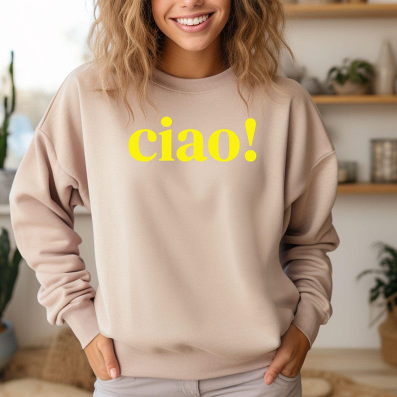 Könnte beinhalten: Ein hellbeiger Pullover mit einem gelben Aufdruck, der "ciao!" sagt.