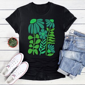 May include: Black t-shirt featuring a green floral design. The design includes stylized flowers and leaves in varying shades of green. The shirt is paired with white sneakers, blue jeans, and a small potted succulent.