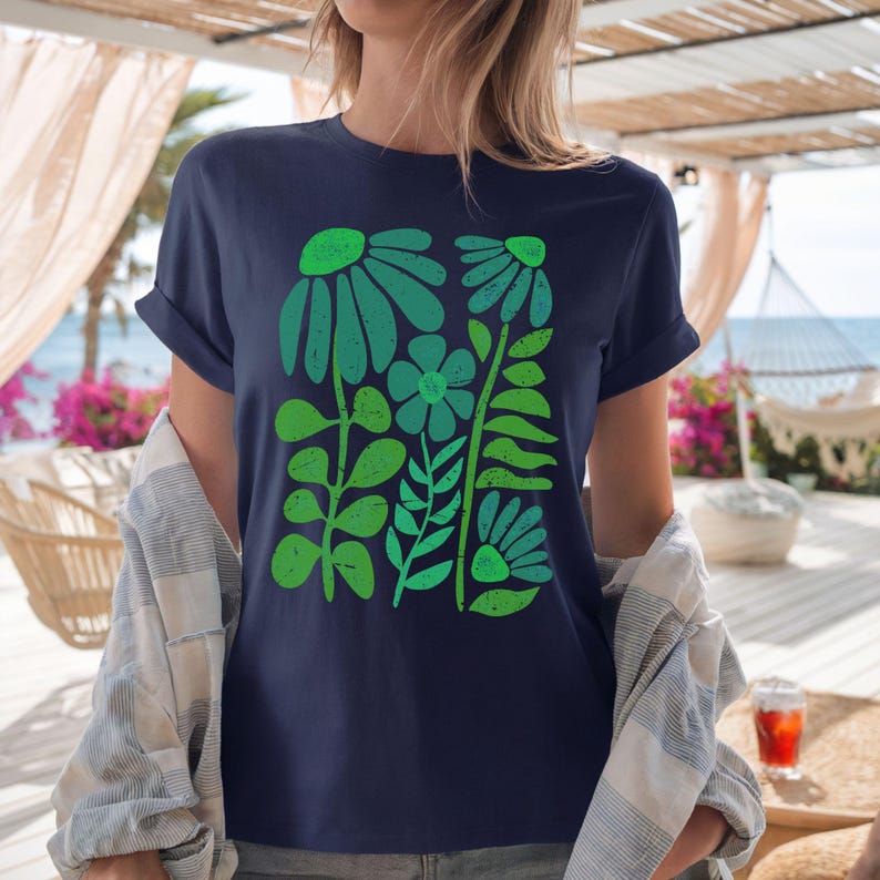 May include: Navy blue t-shirt featuring a green floral design. The graphic includes stylized flowers and leaves in shades of green. The shirt has rolled-up sleeves and is made of a soft material.