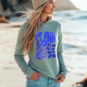 May include: A sage green long-sleeve hooded sweatshirt featuring a central graphic print of stylized blue and purple flowers and leaves. The hoodie has a drawstring hood and ribbed cuffs, paired with light blue denim jeans and a beige knit beanie.