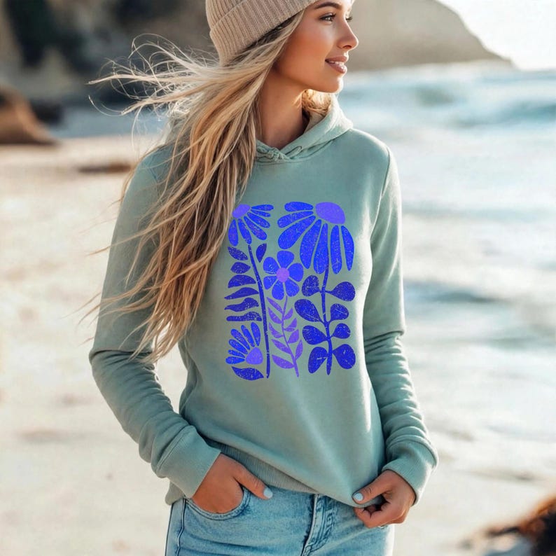 May include: A sage green long-sleeve hooded sweatshirt featuring a central graphic print of stylized blue and purple flowers and leaves. The hoodie has a drawstring hood and ribbed cuffs, paired with light blue denim jeans and a beige knit beanie.