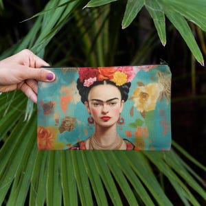 May include: A rectangular cosmetic bag featuring a portrait of Frida Kahlo with a floral headdress. The bag has a teal background with floral accents in orange, yellow, and pink. The zipper is black.
