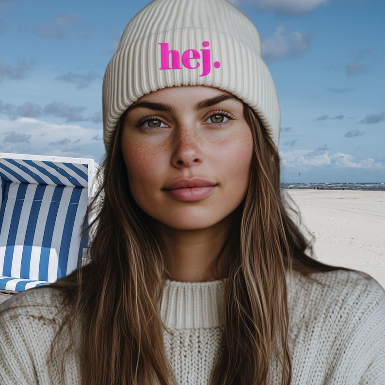 May include: A cream-coloured ribbed knit beanie with the word "hej." in pink. The beanie is worn by a person with long brown hair and a cream-coloured sweater. The background shows a beach scene.