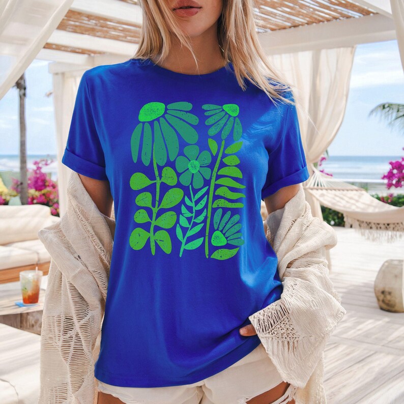 May include: A royal blue t-shirt featuring a green floral design. The graphic includes stylized flowers and leaves in varying shades of green. The shirt has short sleeves and a crew neck.