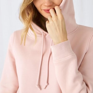 May include: A light pink hooded sweatshirt featuring a matching drawstring with aglets, a hood, and long sleeves with ribbed cuffs. The garment is constructed from a solid-colored fabric with a smooth texture and a clean, simple design.