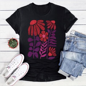 May include: Black t-shirt featuring a floral design in shades of red, pink, and purple. The graphic includes stylized flowers and leaves. The shirt is paired with blue jeans and white sneakers.