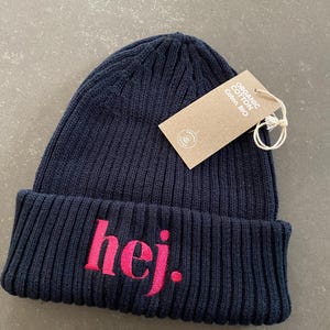 May include: A navy blue ribbed knit beanie with the word "hej" embroidered in pink. A brown tag with the words "Organic Cotton Coton Bio" is attached to the hat.