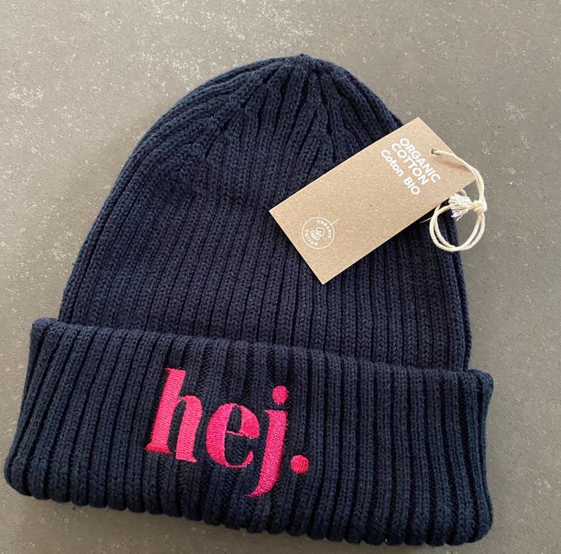 May include: A navy blue ribbed knit beanie with the word "hej" embroidered in pink. A brown tag with the words "Organic Cotton Coton Bio" is attached to the hat.