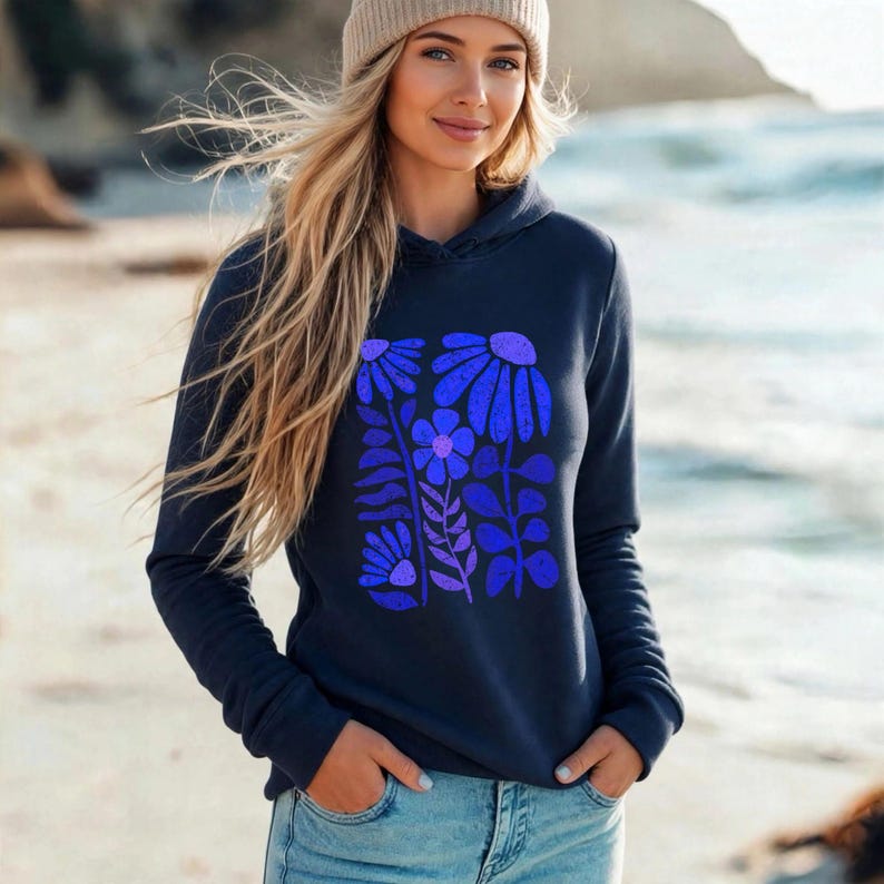 May include: A navy blue hooded sweatshirt featuring a central graphic print of stylized flowers in shades of bright blue and purple. The hoodie has long sleeves, a front kangaroo pocket, and a hood, worn with light blue denim jeans and a beige beanie.