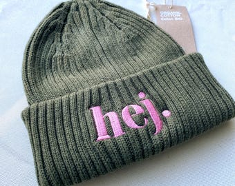 May include: An olive green ribbed knit beanie with a folded cuff. The word "hej." is embroidered in pink thread on the front of the cuff. A brown paper tag attached to the top reads "ORGANIC COTTON Coton BIO" in white text.