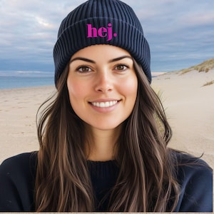 May include: A navy blue ribbed knit beanie with the word "hej." in bright pink lettering. The beanie is worn by a person with long brown hair, smiling in front of a beach scene.