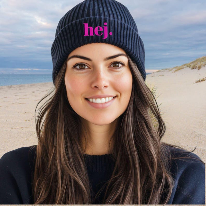 May include: A navy blue ribbed knit beanie with the word "hej." in bright pink lettering. The beanie is worn by a person with long brown hair, smiling in front of a beach scene.