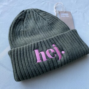 May include: An olive green knitted beanie hat with the word "hej." embroidered in pink. The hat has a ribbed design and a folded brim. A tag reading "Organic Cotton" is attached.