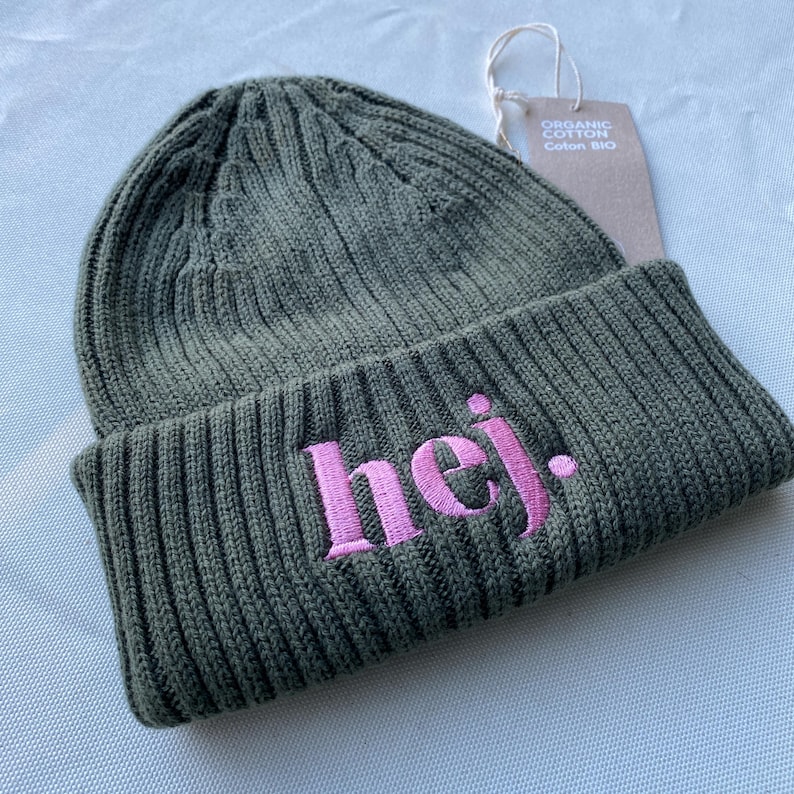 May include: An olive green knitted beanie hat with the word "hej." embroidered in pink. The hat has a ribbed design and a folded brim. A tag reading "Organic Cotton" is attached.