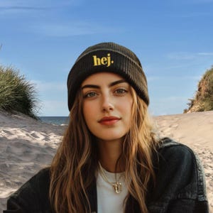 May include: A woman wearing a dark grey beanie hat with the word "hej" in yellow. She is wearing a white t-shirt and a black denim jacket. The background shows a sandy beach and the sea.