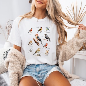 Könnte beinhalten: Weißes T-Shirt mit einer Aquarellillustration verschiedener Vogelarten, die jeweils mit ihrem Namen beschriftet sind. Die Vögel sind in leuchtenden Farben dargestellt und auf Ästen platziert. Das Shirt ist ein lässiges Kleidungsstück.