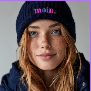 Könnte beinhalten: Eine marineblaue Strickmütze mit dem Wort "moin." in rosa und lila Buchstaben. Die Mütze wird von einer Person mit heller Haut, Sommersprossen und rotem Haar getragen. Die Person trägt eine marineblaue Jacke.