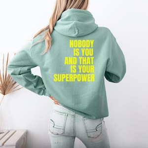 May include: A pale green hoodie with the text "NOBODY IS YOU AND THAT IS YOUR SUPERPOWER" in yellow. The hoodie has a hood and long sleeves. The person is wearing light blue jeans.