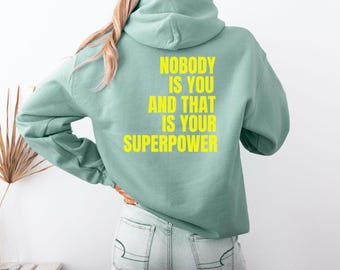 May include: A light green hoodie with the text "NOBODY IS YOU AND THAT IS YOUR SUPERPOWER" in yellow. The hoodie has a hood and long sleeves. The person is wearing light blue jeans.