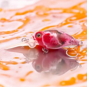 Puede incluir: Un señuelo de pesca rojo y blanco con un labio de plástico transparente y dos anzuelos plateados. El señuelo se refleja en una superficie naranja brillante. La imagen tiene poca profundidad de campo, con el señuelo enfocado.