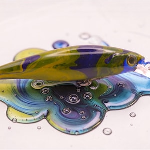 Peut inclure: Un leurre de pêche avec un motif camouflage jaune, vert et bleu. Le leurre a un hameçon argenté et une petite nageoire en plastique transparent. Le leurre repose sur une surface avec un motif tourbillonnant bleu, jaune et vert.