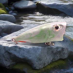 Pode incluir: Um isco de pesca branco e verde com uma faixa vermelha e um anzol prateado. O isco está sobre uma rocha cinza.