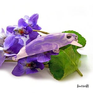 Puede incluir: Un señuelo de pesca morado y blanco con un diseño de mármol. El señuelo está descansando sobre un lecho de flores moradas y hojas verdes.