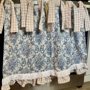 May include: A cafe curtain featuring a blue and white damask pattern on a cream background. The top edge has beige and white gingham tie-top tabs, while the bottom edge is finished with a white eyelet lace ruffle and a gingham fabric trim.