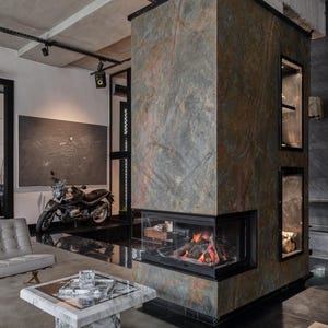 May include: A large, rectangular, multi-sided fireplace column clad in textured, multi-colored stone with gray, brown, and rust tones. The fireplace features a glass-enclosed firebox with burning logs and two recessed vertical display niches.