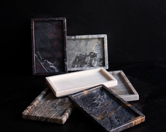 May include: A collection of rectangular marble trays in various colors and patterns. The trays are in shades of black, gray, white, and brown, with veining and marbling details. The trays are arranged on a dark surface.