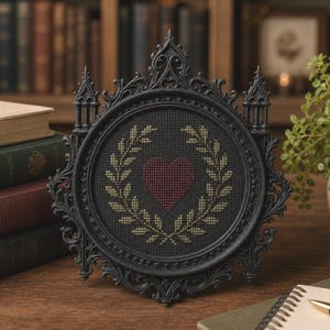 May include: A circular cross-stitch embroidery featuring a dark red heart surrounded by a golden-yellow laurel wreath on a black fabric background, encased in an ornate black frame with pointed architectural details and intricate scrollwork.