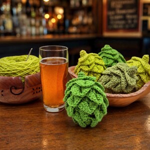 Peut inclure: Un verre transparent de bière ambrée est posé à côté d'un bol à laine en bois contenant une pelote de laine vert clair avec un crochet en métal. Plusieurs ornements en crochet en forme de houblon vert sont disposés dans un bol en bois.