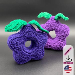 May include: Two handmade crochet toys, one star-shaped and one rounded, both with a central hole. They are a deep purple with green leaf details. The toys are made from a soft, textured yarn, perfect for babies or toddlers.