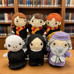 May include: Six crocheted dolls representing fictional characters. The group includes figures with brown, black, and orange yarn hair, one with a lightning bolt, one with a long gray beard, and one bald figure, all wearing various colored outfits.