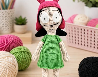 May include: A crocheted doll featuring a character with large white eyes, black hair, and a bright pink bunny-ear hat. The doll wears a simple green dress and black shoes, with light-colored skin tone yarn used for the arms and legs.