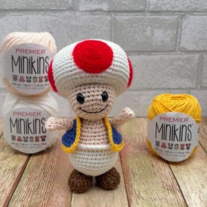 May include: A crocheted Toad character with a red and white mushroom cap, blue vest, and brown shoes. The character is smiling and surrounded by three skeins of Premier Minikins yarn in cream and yellow colors.