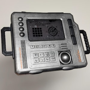 May include: A metallic silver rectangular electronic device with black side handles. The front features a central screen displaying terrain elevation data, a grid, and a radar icon. It includes a numeric keypad, a circular trackball, and various buttons.