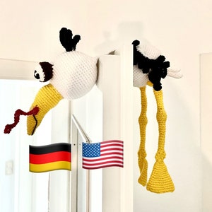 Op de afbeelding: Twee gehaakte knuffels, een witte met een gele snavel en zwarte ogen, en een zwart-witte met gele poten, hangen aan een wit deurkozijn. Een Amerikaanse vlag en een Duitse vlag hangen aan het deurkozijn.