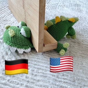 Op de afbeelding: Een groen gehaakt dinosaurus speelgoed met gele accenten, liggend op een wit, getextureerd oppervlak. De dinosaurus is gedeeltelijk verborgen achter een houten plank.