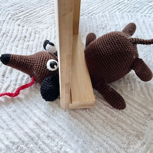 Op de afbeelding: Twee bruine gehaakte hondenspeelgoedjes, een met een rode staart en de andere met een bruine staart, liggen op een wit pluche kleed. De honden zijn gedeeltelijk verborgen achter een houten frame.